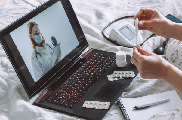 telewizyta lekarska lekarz na ekranie laptopa, dłonie pacjenta trzymające termometr, leki leżące na laptopie