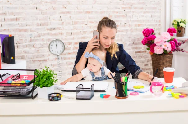 młoda kobieta rozmawiająca przez telefon z niemowlakiem na kolanach siedząca za biurkiem